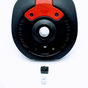 May include: Black and red weight plate components, including a circular main piece with the text "DO NOT DROP", a small black square, a spring, and a red curved piece with two black pegs. The components are arranged on a white background.
