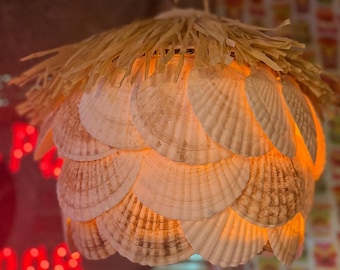 Może przedstawiać: Unikalna lampa wisząca wykonana z muszli, ułożonych w nakładających się warstwach. Lampa ma naturalny, beżowy kolor z górą przypominającą słomę. Światło świeci ciepłym, zachęcającym pomarańczowym odcieniem, tworząc plażową atmosferę.