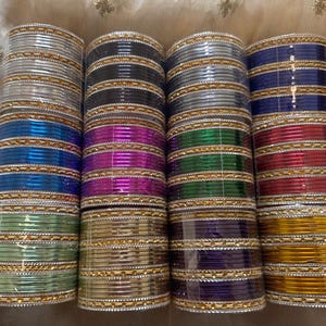 May include: A collection of metal bangles arranged in four vertical stacks. Each stack features alternating bands of solid-colored metallic rings and thin, textured gold-tone spacer rings. The colors include silver, black, blue, light green, purple, red, and gold.