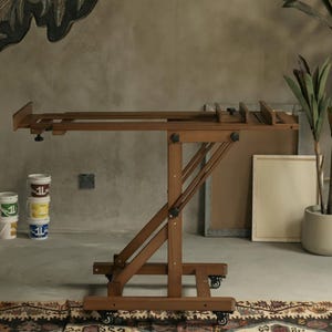 May include: A wooden easel with adjustable height and angle, featuring a brown finish and black hardware. The easel is on wheels and has a canvas holder.