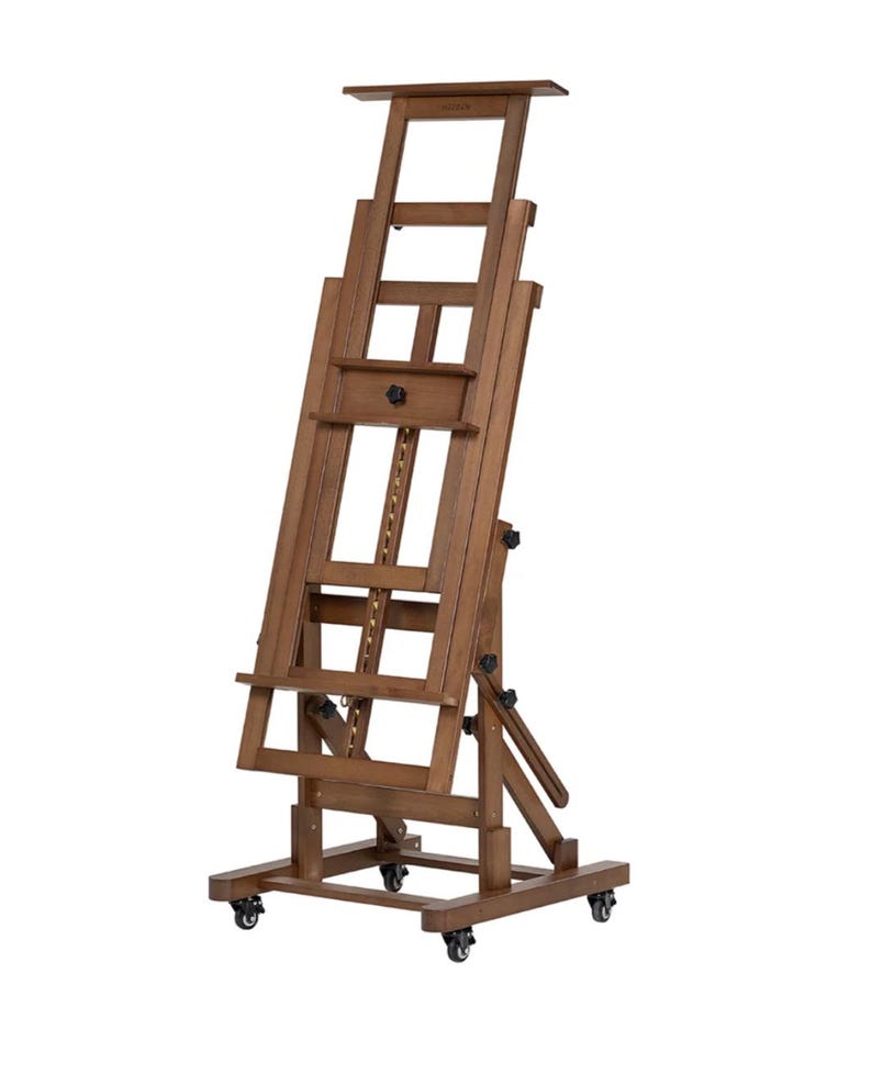 May include: A brown wooden artist's easel with a chain and pulley system for adjusting the height. It features a drawer and a shelf for supplies. The easel is on wheels and can be moved around easily.