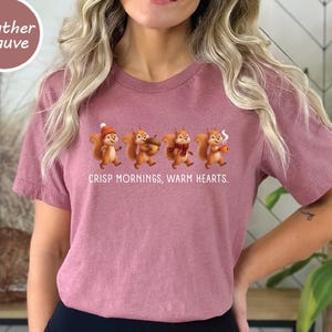May include: A heather mauve t-shirt featuring a graphic of four cartoon squirrels and the text "CRISP MORNINGS, WARM HEARTS." The squirrels are depicted in various poses, with one wearing a hat, one holding an acorn, one wearing a scarf, and one holding a mug.