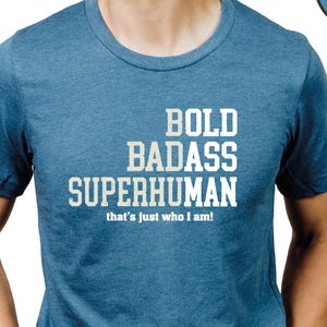 May include: A blue t-shirt with the words "BOLD BADASS SUPERHUMAN" printed in white, with the phrase "that's just who I am!" below. The shirt is a crew neck style.