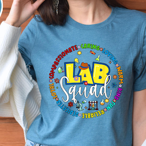 Pode incluir: Uma camiseta azul com as palavras "LAB Squad" em amarelo e branco, rodeada por palavras coloridas e ilustrações com tema científico. A camiseta tem gola redonda e mangas curtas.
