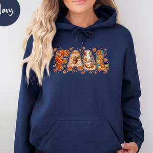 May include: A navy blue hooded sweatshirt featuring a graphic on the chest that spells the word FALL. The letters are filled with autumn-themed patterns including orange leaves, acorns, a coffee mug, and plaid textures, accented by small hearts.