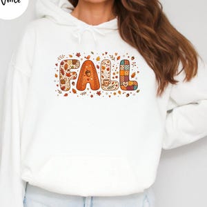 Op de afbeelding: Een witte hoodie met een afbeelding op de borst met het woord FALL in grote, gestileerde letters. Elke letter is gevuld met herfstpatronen, waaronder bladeren, eikels, ruitjes en een dampende mok, omringd door kleine herfsticoontjes.