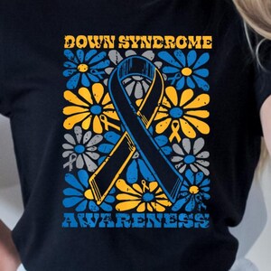 Op de afbeelding: Zwart T-shirt met een ontwerp ter bevordering van het bewustzijn van het syndroom van Down. Het ontwerp toont een blauw en geel lint, omringd door blauwe, gele en grijze bloemen, met de woorden "DOWN SYNDROME AWARENESS" in een retro lettertype.