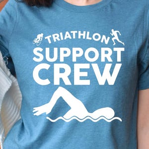 May include: A heather deep teal t-shirt with the words "TRIATHLON SUPPORT CREW" in white. The shirt features white graphic illustrations of a bicycle, a runner, and a swimmer. The shirt is made of a soft material.
