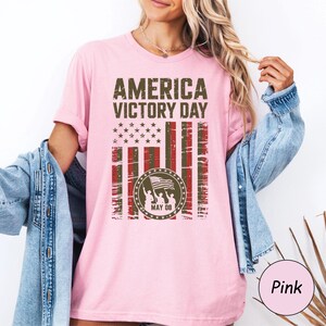 May include: Pink t-shirt with the text "AMERICA VICTORY DAY" in distressed green lettering. The design includes a stylized American flag with stars and stripes, and a circular emblem with figures holding a flag. The date "MAY 08" is also included.