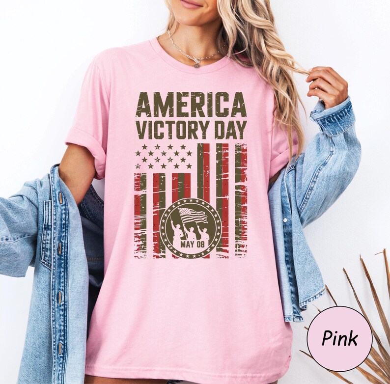 May include: Pink t-shirt with the text "AMERICA VICTORY DAY" in distressed green lettering. The design includes a stylized American flag with stars and stripes, and a circular emblem with figures holding a flag. The date "MAY 08" is also included.