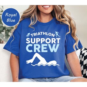 May include: Royal blue t-shirt with the text "TRIATHLON SUPPORT CREW" in white and light blue. The design includes silhouettes of a cyclist, runner, and swimmer. A round label in the upper left corner reads "Royal Blue".