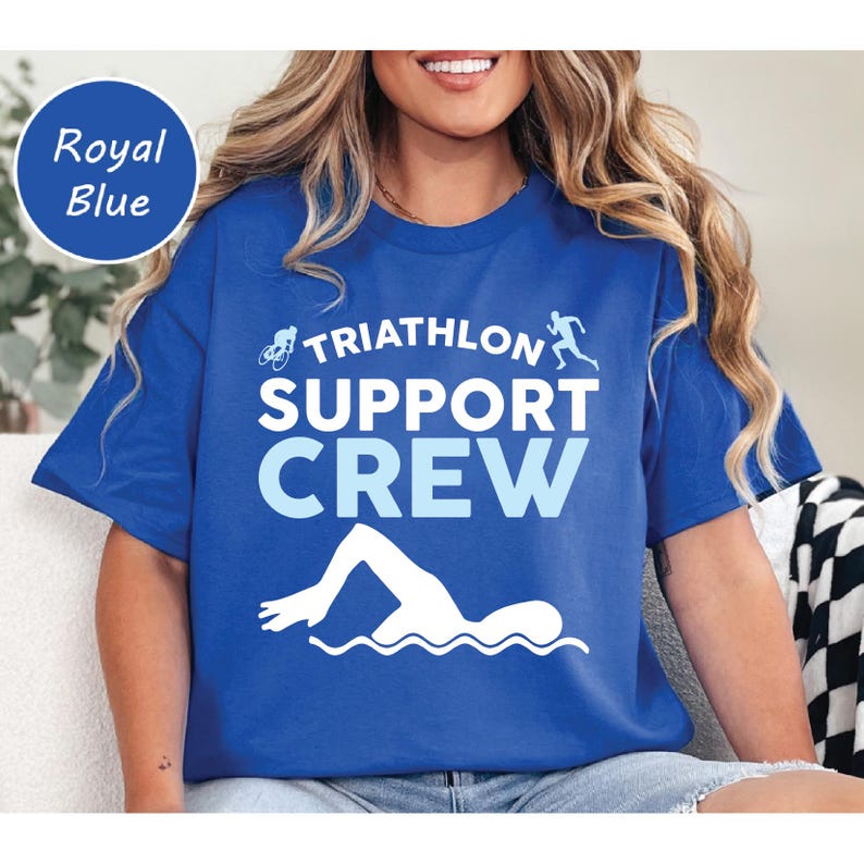 May include: Royal blue t-shirt with the text "TRIATHLON SUPPORT CREW" in white and light blue. The design includes silhouettes of a cyclist, runner, and swimmer. A round label in the upper left corner reads "Royal Blue".