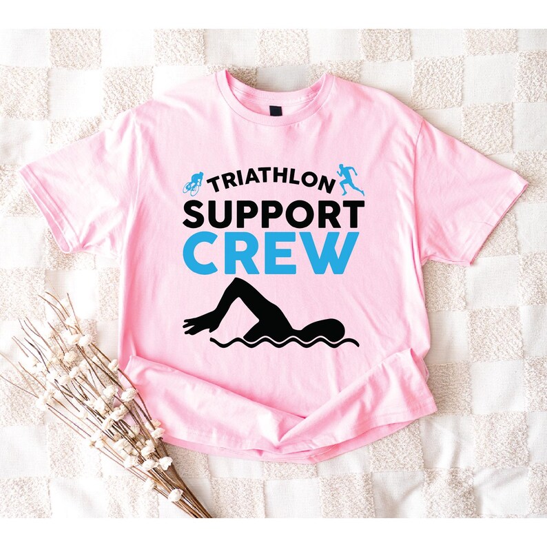 May include: A light pink t-shirt with the words "TRIATHLON SUPPORT CREW" in black and blue. The shirt features a swimmer graphic and icons of a bicycle and runner. The shirt is laid flat on a white and light pink checkered surface.