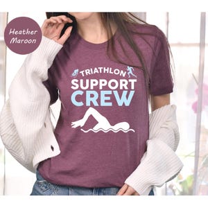 May include: Heather maroon t-shirt with the text "TRIATHLON SUPPORT CREW" in white and light blue. The design includes a swimmer, a cyclist, and a runner. The shirt is made of a soft material.