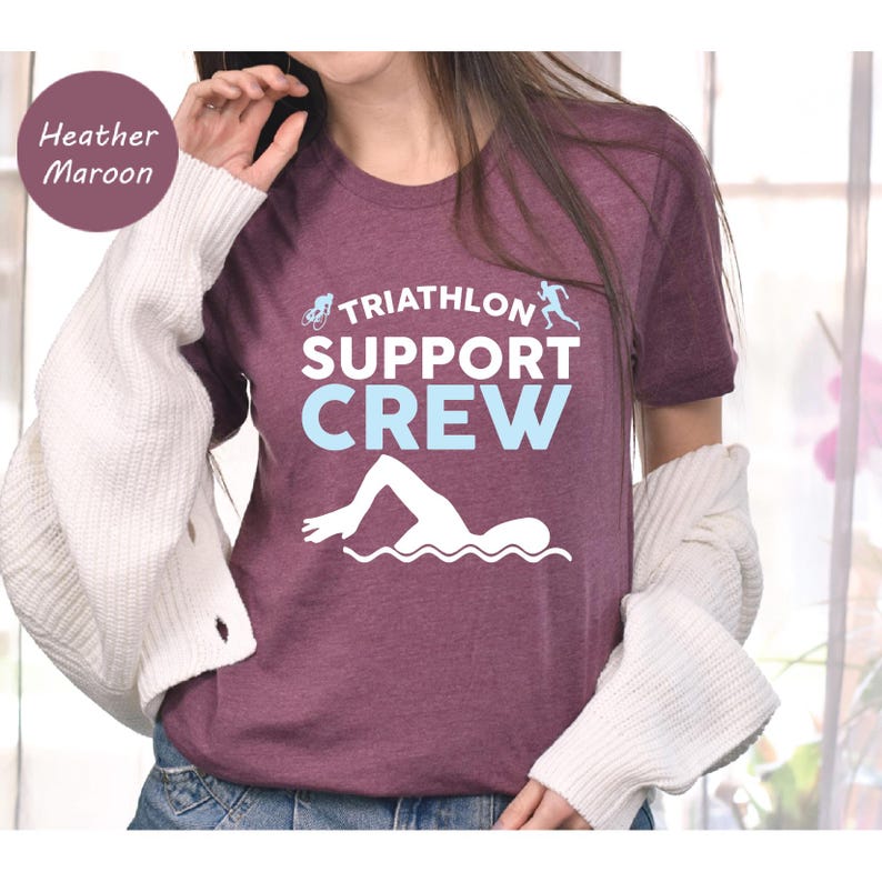 May include: Heather maroon t-shirt with the text "TRIATHLON SUPPORT CREW" in white and light blue. The design includes a swimmer, a cyclist, and a runner. The shirt is made of a soft material.