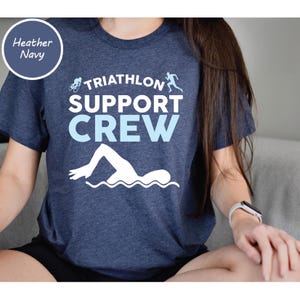 May include: Heather navy blue t-shirt with the words "TRIATHLON SUPPORT CREW" in white and light blue. The shirt features a swimmer, a cyclist, and a runner graphic. The shirt is made of a soft material.