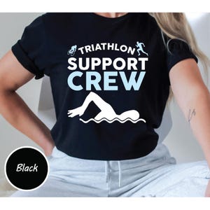 May include: Black t-shirt with the text "TRIATHLON SUPPORT CREW" in white and light blue. The design includes a cyclist, a runner, and a swimmer. A black circle with the word "Black" is in the lower left corner.