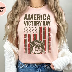 May include: A light pink t-shirt with the text "AMERICA VICTORY DAY" in dark brown. The design includes a distressed American flag graphic and a circular emblem with figures holding a flag, with "MAY 08" below.