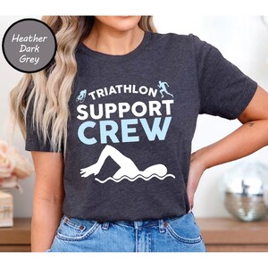 May include: Heather dark grey t-shirt with the words "Triathlon Support Crew" in white and light blue. The shirt features illustrations of a cyclist, runner, and swimmer. The shirt is made of a soft material.