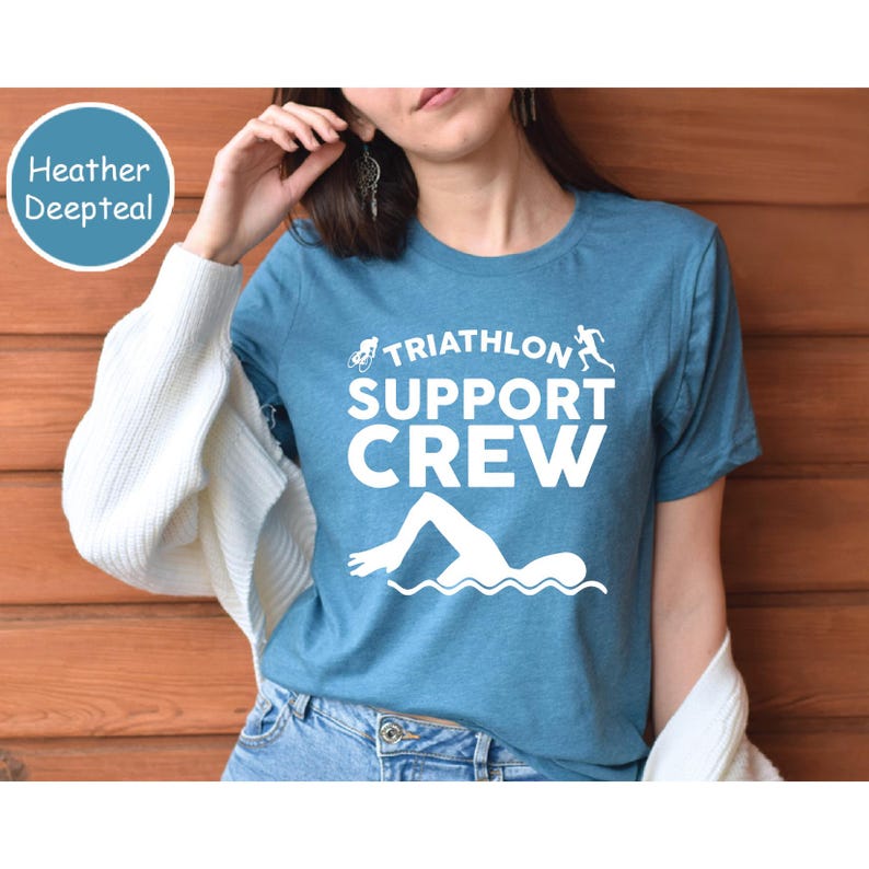 May include: A heather deep teal t-shirt with the words "TRIATHLON SUPPORT CREW" in white. The shirt features white graphic illustrations of a bicycle, a runner, and a swimmer. The shirt is made of a soft material.