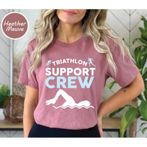 May include: Heather mauve t-shirt with the text "TRIATHLON SUPPORT CREW" in white and light blue. The shirt features illustrations of a cyclist, runner, and swimmer. A round label in the upper left corner reads "Heather Mauve".