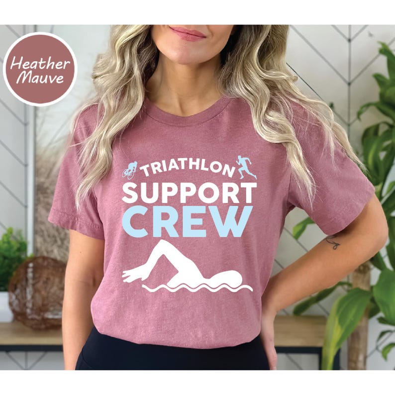 May include: Heather mauve t-shirt with the text "TRIATHLON SUPPORT CREW" in white and light blue. The shirt features illustrations of a cyclist, runner, and swimmer. A round label in the upper left corner reads "Heather Mauve".