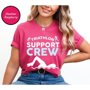 May include: Heather raspberry t-shirt with the text "TRIATHLON SUPPORT CREW" in white and light blue. The design includes a bicycle, a runner, and a swimmer. The shirt is a crew neck and has rolled-up sleeves.
