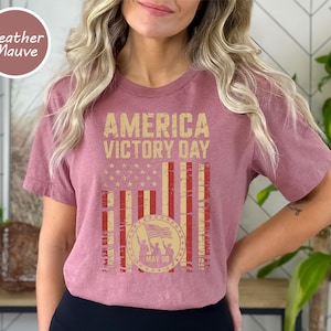 May include: Heather mauve t-shirt with a distressed graphic design. The design features the text "AMERICA VICTORY DAY" above a stylized American flag with a circular emblem. The emblem includes a waving flag and the date "MAY 08".