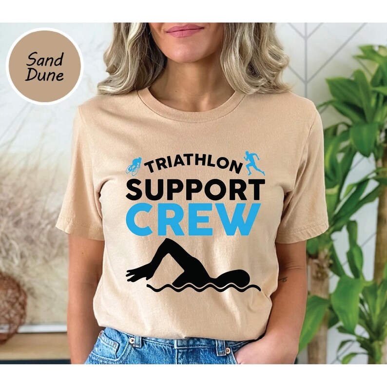 May include: A tan t-shirt with the words "TRIATHLON SUPPORT CREW" in black and blue. The design includes silhouettes of a cyclist, runner, and swimmer. The shirt is made of a soft material.