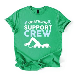 May include: A green t-shirt with the words "TRIATHLON SUPPORT CREW" in white and light blue. The shirt features white graphics of a cyclist, runner, and swimmer. The t-shirt is made of a soft material.