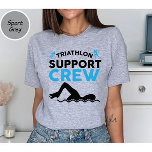 May include: Sport grey t-shirt with the words "TRIATHLON SUPPORT CREW" in black and blue. A black swimmer graphic is below the text. The shirt is a crew neck and short-sleeved.