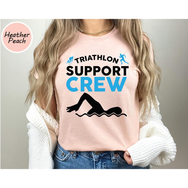 May include: A light pink t-shirt with the words "TRIATHLON SUPPORT CREW" in black and blue. A black silhouette of a swimmer is below the text. The shirt also has the words "Heather Peach" in a circle.
