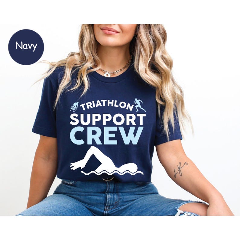 May include: Navy blue t-shirt with the words "TRIATHLON SUPPORT CREW" in white and light blue. The shirt features illustrations of a cyclist, runner, and swimmer. The shirt is made of a soft material.
