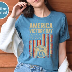 May include: A heather deepteal t-shirt with the words "AMERICA VICTORY DAY" in a distressed gold font. The design includes a flag graphic with red and white stripes, gold stars, and a circular emblem with the date "MAY 08".
