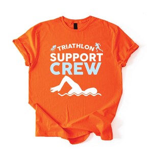 May include: Orange t-shirt with the words "TRIATHLON SUPPORT CREW" in white and light blue. The shirt features a swimmer graphic, a runner graphic, and a bicycle graphic. The shirt is made of a soft material.