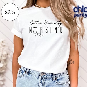 May include: White t-shirt with the text "Custom University Nursing" in black script and a stethoscope graphic. The shirt has short sleeves and is worn by a person in blue jeans.