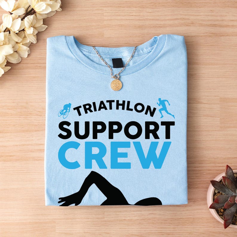 May include: A light blue t-shirt with the words "TRIATHLON SUPPORT CREW" in black and blue. The shirt features illustrations of a cyclist, runner, and swimmer. A gold necklace with a round pendant is on the shirt.