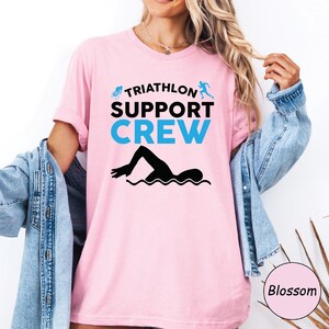May include: A light pink t-shirt with the words "TRIATHLON SUPPORT CREW" in black and blue. A black silhouette of a swimmer is below the text. The shirt has short sleeves.