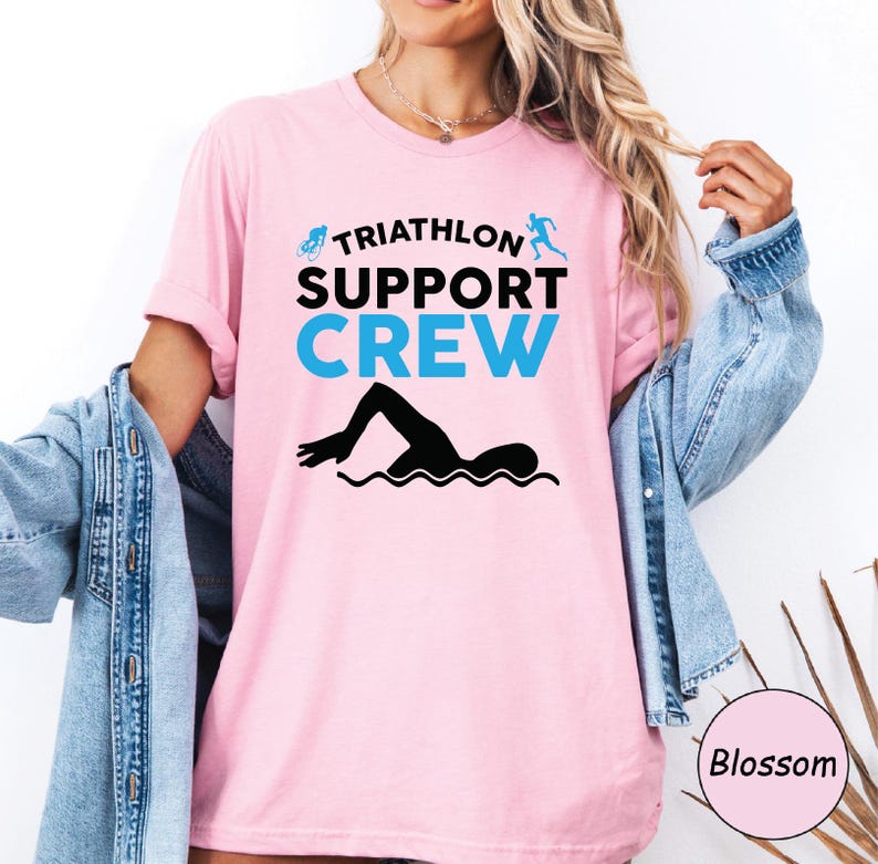 May include: A light pink t-shirt with the words "TRIATHLON SUPPORT CREW" in black and blue. A black silhouette of a swimmer is below the text. The shirt has short sleeves.