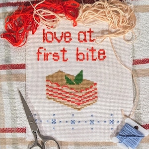 May include: A white cross-stitch embroidery with the text "love at first bite" and a depiction of a layered dessert with green leaves on top. The embroidery is surrounded by red, orange, and blue embroidery floss.