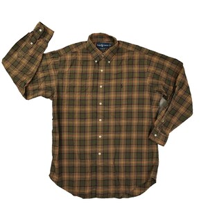 May include: A button-down plaid shirt in shades of olive green, brown, and orange. The long-sleeved shirt has a button-down collar and a small embroidered logo on the chest. The shirt is laid flat on a white surface.