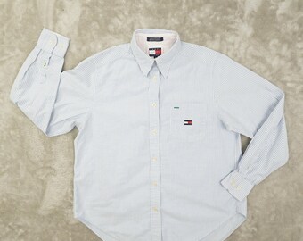 May include: A long-sleeve button-down shirt featuring a light blue and white vertical pinstripe pattern. The shirt includes a pointed collar, a single chest pocket with a small embroidered flag logo, and buttoned cuffs on the long sleeves.