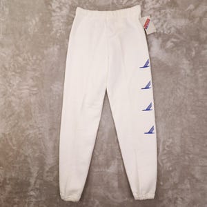 May include: White sweatpants with an elastic waistband and cuffs. The pants feature four blue stylized wing designs down the left leg. A tag is attached to the waistband. The pants are made of a soft material.
