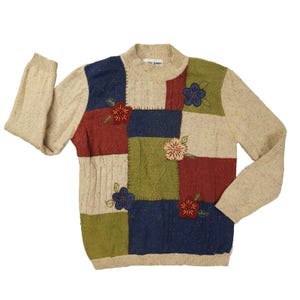 May include: A long-sleeve sweater featuring a patchwork design of beige, navy blue, olive green, and dark red panels. The sweater includes cable knit textures and embroidered floral appliqués in red, blue, and cream. It has a mock neck collar.