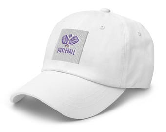 Puede incluir: Gorra de béisbol blanca con un logotipo de pickleball bordado en morado. El logotipo presenta dos palas y una pelota.