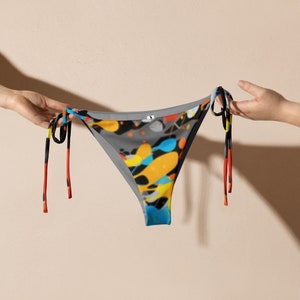 May include: A bikini bottom with a gray background and colorful abstract paint splatters in yellow, blue, orange, and black. The bikini has black ties with red, yellow, and black stripes. The bikini is held by two hands.