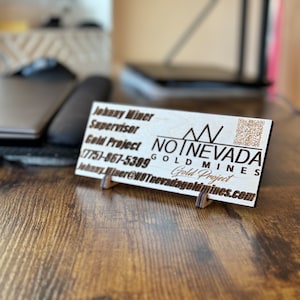 Könnte beinhalten: Ein Visitenkartenhalter aus Holz mit weißem Hintergrund und schwarzem Text. Der Text lautet "Johnny Miner, Supervisor, Goldprojekt, (775)-867-5399, Johnny Miner@HOTnevadagoldmines.com". Die Karte zeigt auch ein Logo mit dem Text "NO NEVADA GOLD MINES, Goldprojekt".