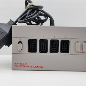 Puede incluir: Adaptador Nintendo NES FOUR SCORE gris claro con cuatro puertos de controlador, dos botones turbo y un cable de alimentación negro. El texto "Nintendo NES FOUR SCORE" está impreso en rojo.