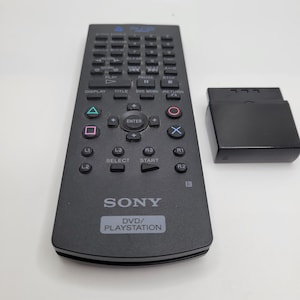 May include: A black Sony DVD/PlayStation remote control with multiple buttons, including play, pause, and directional controls. The remote is accompanied by a black adapter. The text "SONY DVD/PLAYSTATION" is visible on the remote.