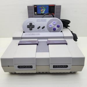 Peut inclure: Une console Super Nintendo Entertainment System gris clair avec une manette assortie posée dessus. Une cartouche de jeu Super Mario World est insérée dans la fente arrière. La console dispose d'un interrupteur, d'un bouton reset et deux ports.